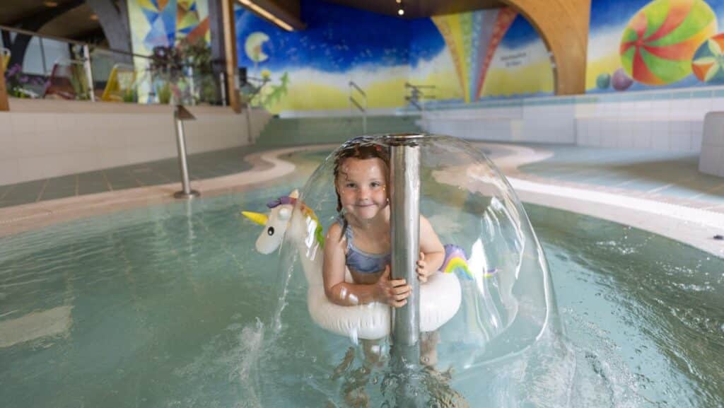 Kleines Mädchen unter Wasserpilz mit Einhaornschwimmreifen im Babybecken des Zzguspitzbades Grainau