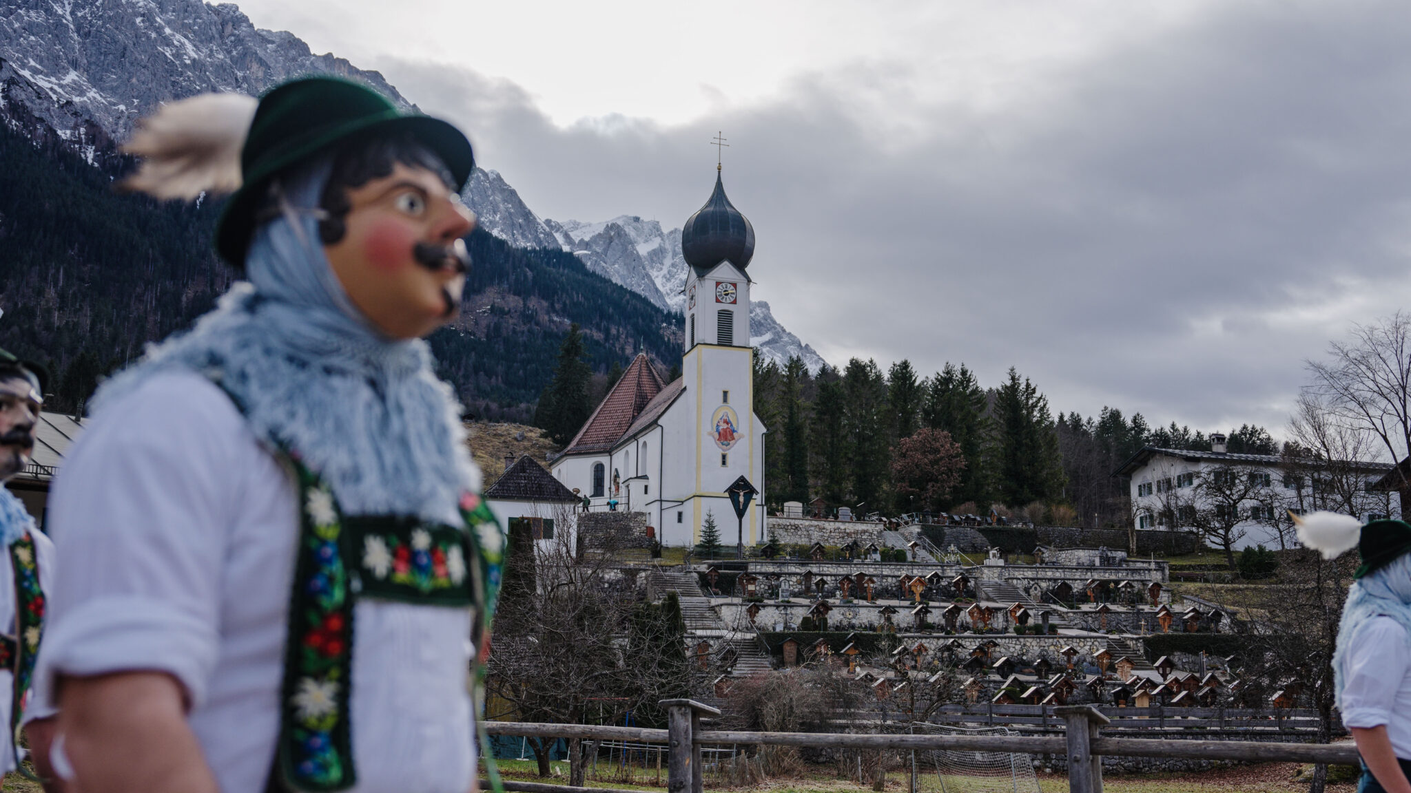 Maschkera dahinter Kirche und Friedhof mit bergen
