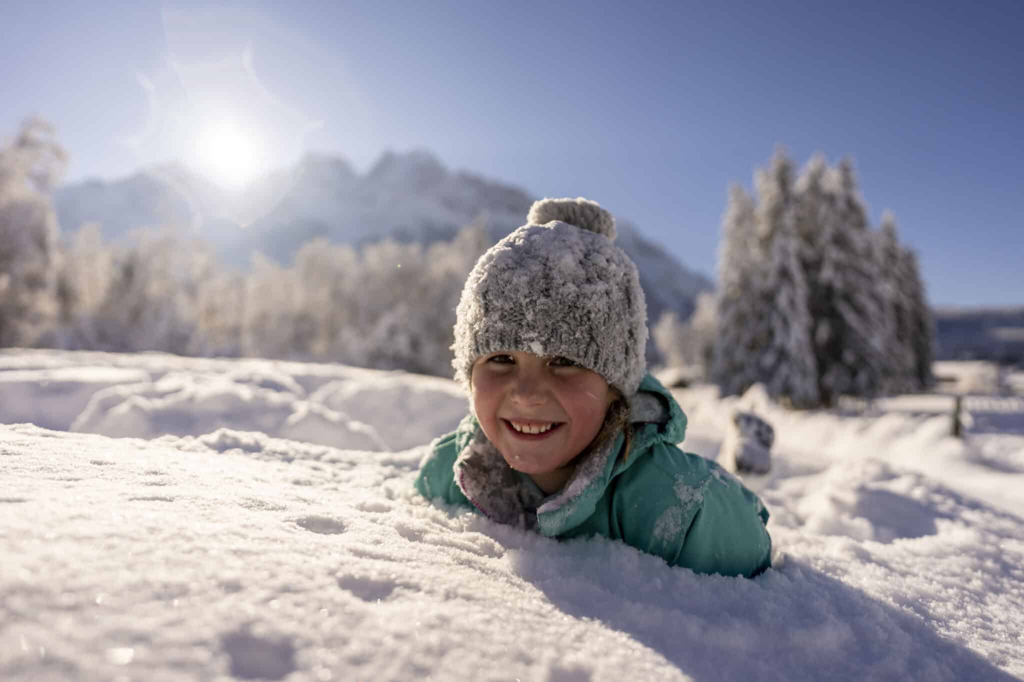Kind im Schnee, nur Kopf sieht aus tiefen Schnee, dahinter verschwommene Zugspitzmassiv und Sonne