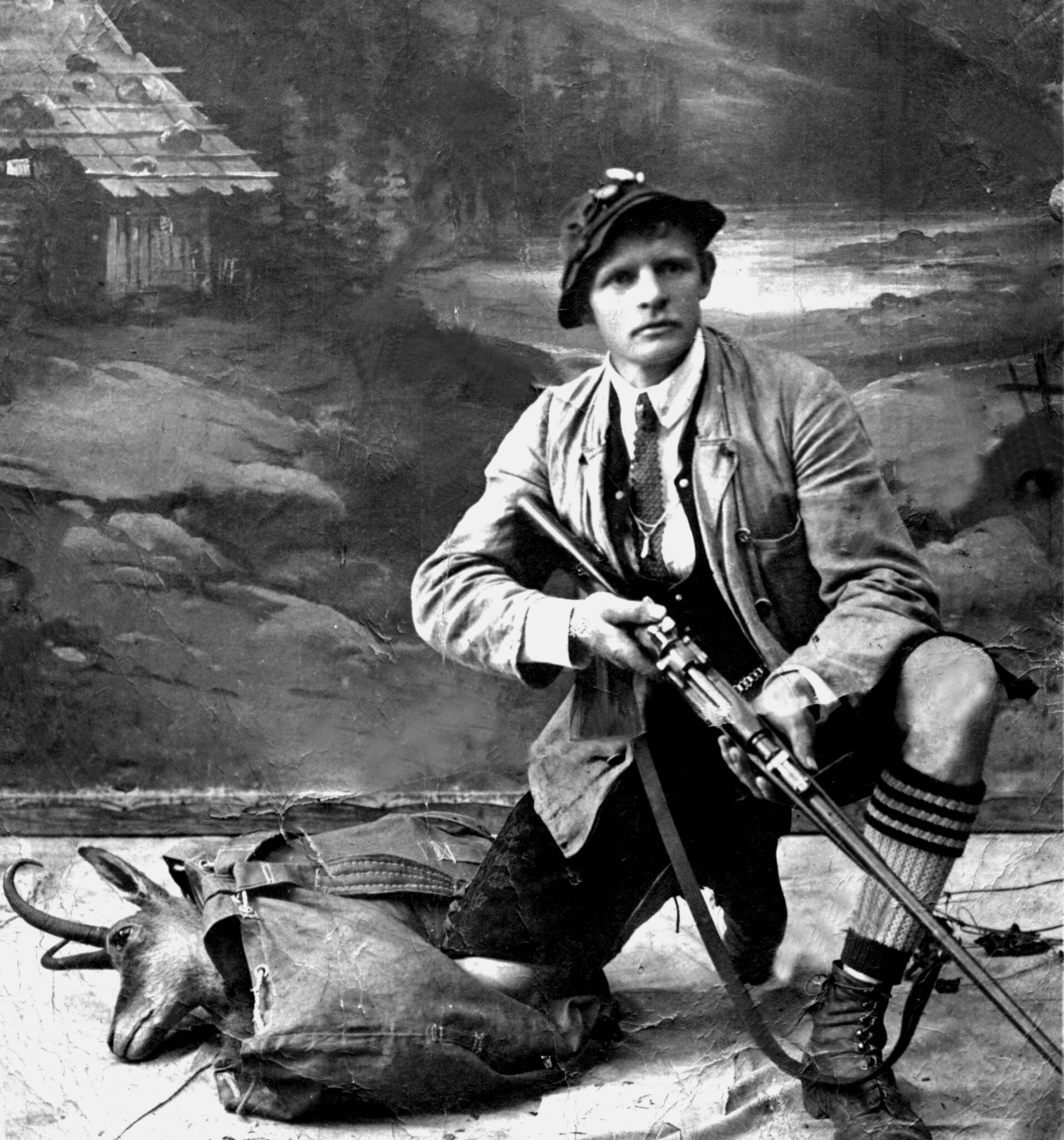 historischer Wilderer mit Stutzen in der Hand knieend, daneben ein Rucksack mit Gamskopf.